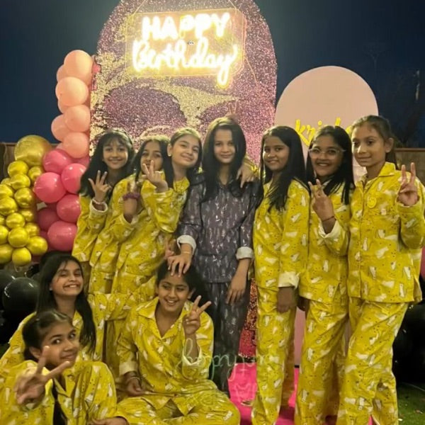 Group of children in yellow matching pajamas posing in front of a 'Happy Birthday' sign with balloons.