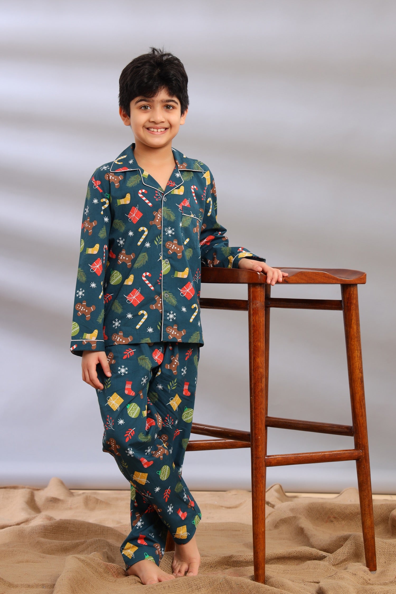Boy wearing 100% cotton Christmas print pajama set standing next to a wooden stool