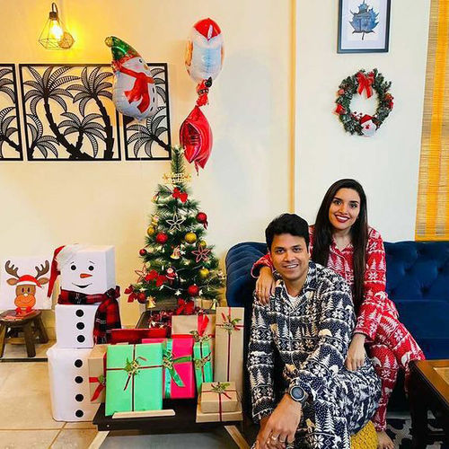 Couple sitting in a living room wearing Christmas pajamas decorated for Christmas with a small tree, presents, and festive balloons