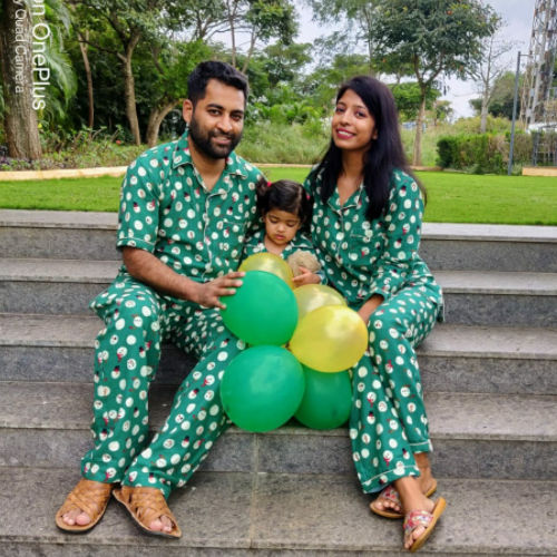 Family of three in Christmas matching green Santa print nightwear with balloons sitting on steps outdoors.