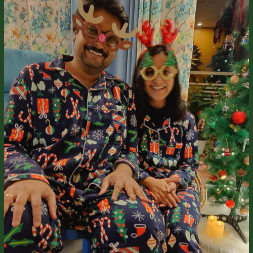 Couple wearing festive Christmas pajamas and reindeer antlers in front of a decorated Christmas tree.