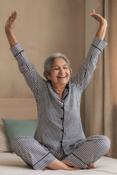 Senior Woman in striped pajamas sitting on a bed with arms raised, smiling.