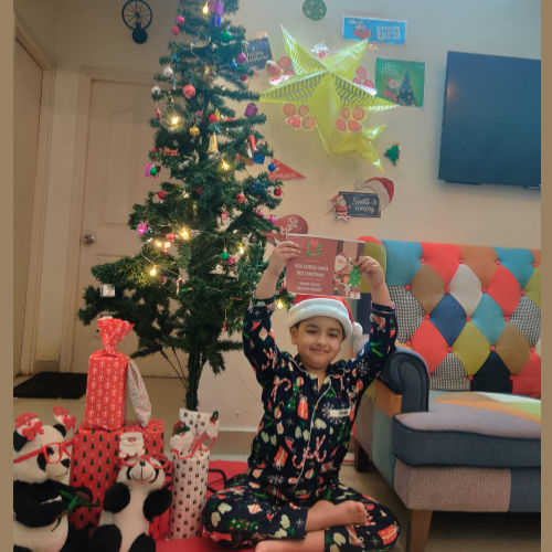 Child in Christmas pajamas holding a card in front of a decorated Christmas tree and colorful chair.